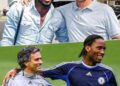 A photo of three Chelsea legends — Michael Essien, Jose Mourinho, and Didier Drogba — standing together during their time at Chelsea. Mourinho is in a suit, while Essien and Drogba wear Chelsea training kit. All three are smiling, celebrating a victory.