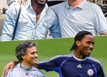 A photo of three Chelsea legends — Michael Essien, Jose Mourinho, and Didier Drogba — standing together during their time at Chelsea. Mourinho is in a suit, while Essien and Drogba wear Chelsea training kit. All three are smiling, celebrating a victory.